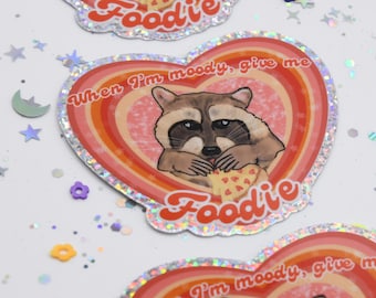 May include: A heart-shaped sticker with a raccoon eating pizza. The sticker has a rainbow design and the text "When I'm moody, give me Foodie".