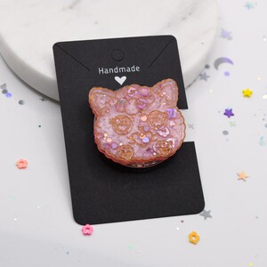 May include: A pink glitter cat face resin charm with the word "Handmade" on a black card.