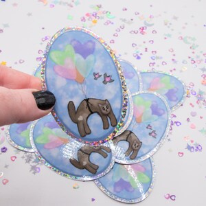 May include: A set of iridescent oval stickers featuring a cartoon cat holding onto a bunch of pastel heart-shaped balloons. The cat is flying in a blue sky with clouds.