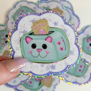 May include: A light blue toaster with a cat face and pink hearts on the side. The toaster has a paw print on top and is surrounded by white stars on a light blue background. The sticker has a holographic glitter border.