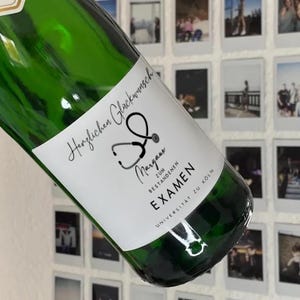 Puede incluir: Una botella de vidrio verde con una etiqueta blanca. La etiqueta presenta el texto "Herzlichen Glückwunsch" y un gráfico de estetoscopio. Debajo, el nombre "Margaux" y la palabra "EXAMEN". La botella es probablemente un regalo de celebración.