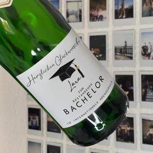Op de afbeelding: Een groene glazen fles met een wit etiket. Het etiket toont een afstudeerhoed, de naam "Lara" en het woord "Bachelor". De fles is waarschijnlijk een cadeau voor een afgestudeerde.