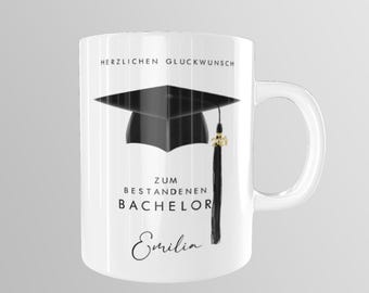 May include: White ceramic mug featuring a black graduation cap and tassel design. The text "Herzlichen Glückwunsch zum bestandenen Bachelor Emilia" is printed on the mug.