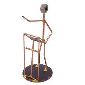May include: A copper and brass sculpture of a person standing at a podium. The person is made of wire and has a nut for a head. The podium is made of wire and has a flat top.