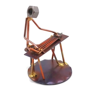 May include: A metal sculpture of a person playing a musical instrument. The sculpture is made of copper and brass and features a nut for a head. The person is sitting on a stool and playing a long, narrow instrument with multiple strings.