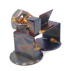 May include: A metal sculpture of a person sitting at a desk with a laptop. The sculpture is made of recycled metal and has a rustic, industrial look. The person is sitting in a chair with their legs crossed. The laptop is open and the screen is facing the viewer. The sculpture is painted in shades of blue, gold, and silver.