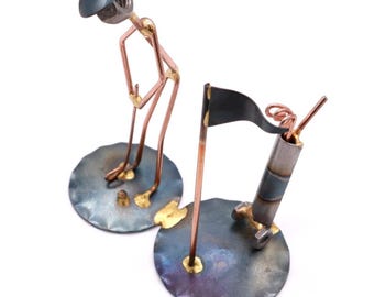 May include: A metal sculpture of a golfer putting a golf ball into a hole. The sculpture is made of various metal pieces, including nuts, bolts, and wire. The golfer is standing on a round metal base, and the hole is on a separate round metal base. The sculpture is painted in a variety of colors, including black, silver, and gold.
