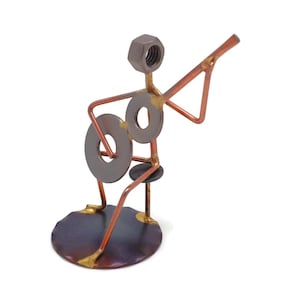 May include: A small metal sculpture of a person playing a guitar. The sculpture is made of various metal pieces, including nuts, bolts, and washers. The person is sitting on a stool and holding a guitar made of wire and washers.
