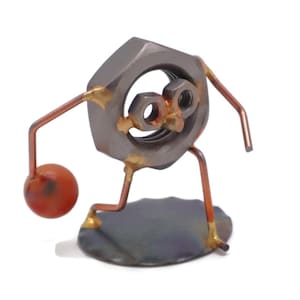 May include: A small metal sculpture of a person playing basketball. The person is made of nuts and bolts, and the basketball is a small orange ball. The sculpture is standing on a metal base.