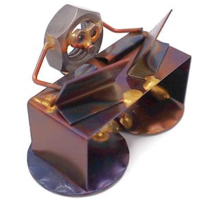 May include: A handcrafted metal sculpture of a person seated at a desk. The figure's head is a large nut, with wire arms and a body made of folded metal. The sculpture has a mix of bronze, purple, and gold tones.