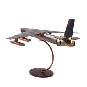 May include: A metal model of a B-52 bomber airplane with a gray and white striped fuselage, copper colored landing gear, and a gold colored engine. The model is mounted on a copper colored stand.
