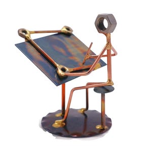 May include: A metal sculpture of a person sitting at a desk, drawing. The person is made of copper wire and nuts, and the desk is made of metal. The sculpture is on a round base.