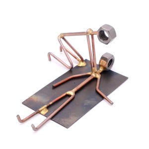 May include: A metal sculpture of two figures, one lying down and the other standing over them, with their heads made of nuts. The figures are welded onto a metal base.