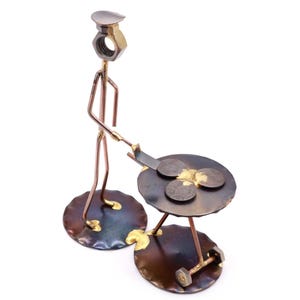 May include: A metal sculpture of a person flipping a pancake on a small grill. The sculpture is made of copper and brass and has a rustic look.