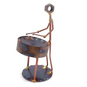 May include: A metal sculpture of a person playing a steel drum. The sculpture is made of various metal pieces, including nuts, bolts, and wire. The steel drum is a dark brown color with a metallic sheen. The person is holding two drumsticks in their hands.