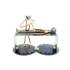 May include: A metal sculpture of two figures, one sitting and the other lying down, on a black metal base. The figures are made of copper wire and nuts, and the base has a gold accent.