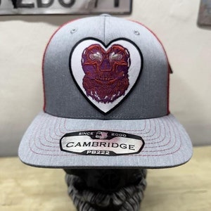 May include: Gray and red baseball cap with a heart-shaped patch featuring a purple and red design. The cap has a flat brim and the text "CAMBRIDGE" is visible on the brim. The cap is a PB222 model.