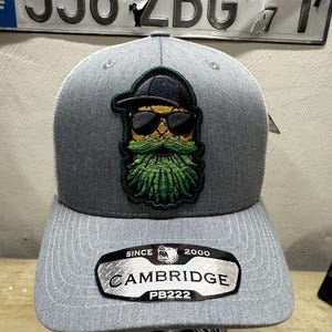 May include: Gray and white trucker hat with a green and yellow bearded man wearing sunglasses and a black hat patch. The hat has a silver patch that says "SINCE 2000 CAMBRIDGE PB222".