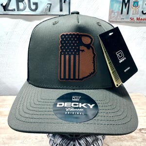 May include: A green trucker hat with a brown leather patch featuring an American flag and a beard. The hat has a black mesh back and a black label with the text "DECKY Classic ORIGINAL".