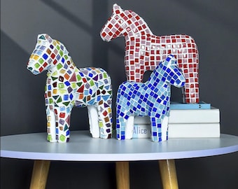 May include: Three horse figurines decorated with colorful mosaic tiles. One horse features a multi-colored pattern, another is covered in red tiles, and the third is adorned with blue tiles. The figurines are displayed on top of white books.