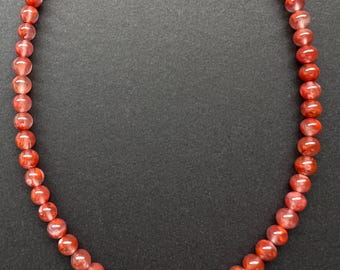 May include: A strand of red gemstone beads, with a subtle shimmer, strung on a thin cord. The beads are round and smooth, and the necklace is approximately 18 inches long.