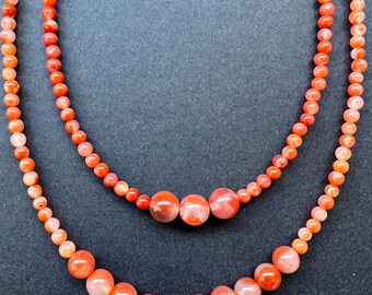 May include: Two strands of orange carnelian beads, one strand is larger than the other, both strands are on a black background.