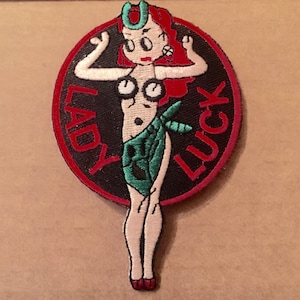 May include: Embroidered patch featuring a cartoon woman with red hair and a green outfit. The patch is circular with the words "LADY LUCK" in red lettering on a black background, surrounded by a red border.