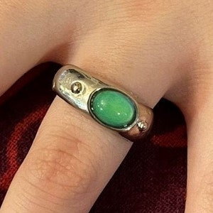 May include: A silver ring with a green oval-shaped stone. The ring has a textured band and a small, round accent stone. The ring is worn on a finger.