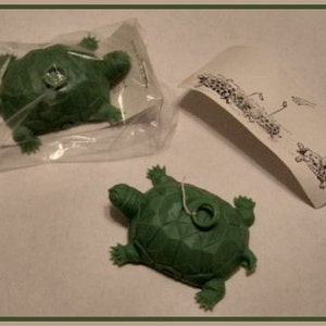 May include: Two green plastic toy turtles. One turtle is in a plastic bag and the other is on a white surface. The turtle on the surface has a string attached to it.