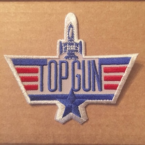 May include: Embroidered Top Gun patch featuring the film's logo in blue and red, with a white outline. The patch has a star and a stylized aircraft design. The patch is on a brown background.