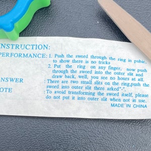 May include: A white card with blue text explaining how to perform a magic trick with a sword and a ring. The text reads "INSTRUCTION: PERFORMANCE: 1. Push the sword through the ring in public, to show there is no tricks 2. Put the ring on any finger, now push through the sword into the outer slit and draw back, well, you see no hours at all. ANSWER: There are two small slits on the ring,push the sword into outer slit there arked"-" To avoid transforming the sword itself, please do not put it into outer slit when not in use. MADE IN CHINA"