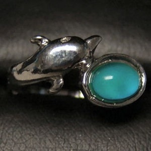 May include: Silver-toned ring featuring a dolphin design. The ring has a turquoise-colored, oval-shaped stone set in a bezel. The dolphin's body curves around the finger, with the stone positioned at the front.