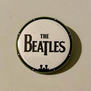 May include: A round button with a white center and a green border. The button features the black text "THE BEATLES" in a bold, sans-serif font. The button has a black rim with small white dots.