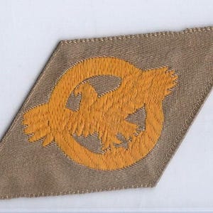 May include: A tan-colored patch with a yellow embroidered design of an eagle inside a circle. The patch has a diamond shape with a tan border and a yellow embroidered eagle.
