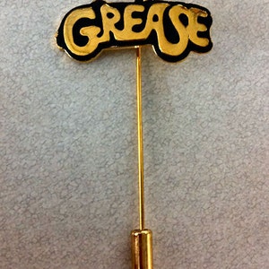 May include: A gold-colored pin with a black outline featuring the word "Grease" in a stylized font. The pin is attached to a long, thin, gold-colored stick.
