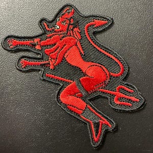 May include: Embroidered patch featuring a red devil woman with horns, a tail, and a trident. The devil figure is set against a black background with a textured appearance. The patch has a black border.