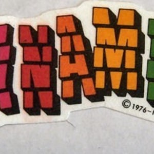 May include: A vintage sticker with the word "DYNAMITE" in colorful, block letters. The letters transition from dark purple to red, orange, green, blue, and purple. The sticker has a white background and the copyright information "© 1976-NADEL S.T.C.-N.Y.C."