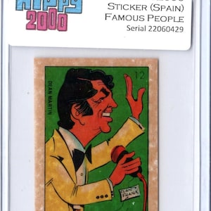 May include: A vintage trading card featuring a cartoon illustration of Dean Martin. The card is from the 1973 HIPPY2000 Famous People series, made in Spain. The card is encased in a protective holder. The card has a green background and the text "CLAN FRANK".