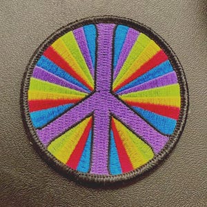 May include: Embroidered patch featuring a vibrant peace symbol. The design includes a purple peace sign at the center, surrounded by colorful rays in shades of red, orange, yellow, green, blue, and purple. The patch has a black border.