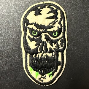 May include: Embroidered patch featuring a menacing skull design. The skull has black outlines and details, with glowing green eyes and accents. The patch is on a cream-colored background, with a jagged edge.