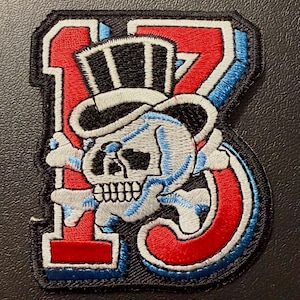 May include: Embroidered patch featuring the number 13 in red, outlined in blue and white, with a skull wearing a top hat and crossed bones. The skull is light blue and gray, with black details. The patch has a black border.