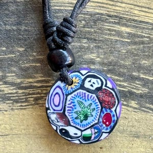 May include: A round pendant necklace with a black cord. The pendant features a colorful mosaic design with various shapes and symbols, including a green leaf, yin-yang, and other geometric patterns. The pendant is approximately 1.5 inches in diameter.