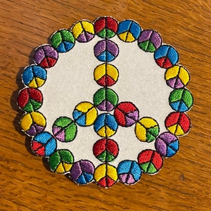 May include: Embroidered peace sign patch with a white background and scalloped edge. The peace symbol is composed of colorful segments in red, yellow, green, blue, and purple. The patch is likely for clothing or accessories.
