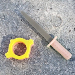 May include: A pink and gray plastic toy sword with a yellow plastic ring on a gray background.