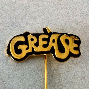 May include: A gold-colored pin with a black outline of the word "Grease" in a stylized font. The pin is attached to a gold-colored pin back.