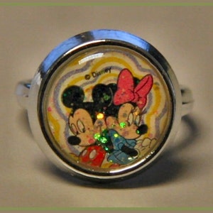 May include: A silver-toned ring featuring Mickey and Minnie Mouse. The ring's centerpiece showcases the iconic Disney characters against a swirling, colorful background. The ring is round and has a shiny, metallic band.