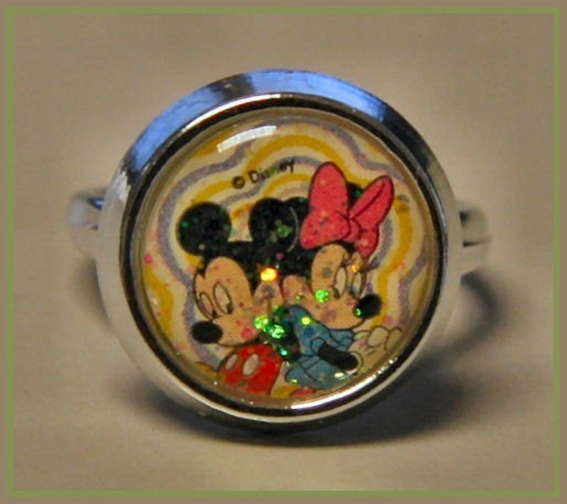May include: A silver-toned ring featuring Mickey and Minnie Mouse. The ring's centerpiece showcases the iconic Disney characters against a swirling, colorful background. The ring is round and has a shiny, metallic band.