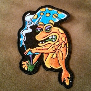 May include: An orange and yellow frog wearing a blue mushroom hat with white spots. The frog has a green mouth and is holding a purple mushroom. The frog is embroidered on a black background.