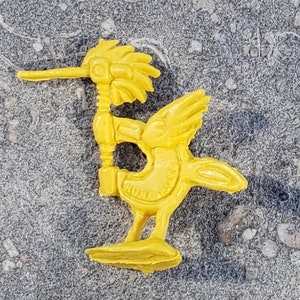 May include: Yellow plastic toy bird with a long beak and a crest on its head. The bird is standing on two legs and has the words "Hong Kong" printed on its body.