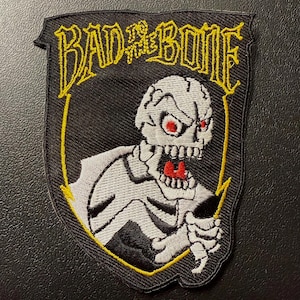 May include: Black patch with a shield shape, featuring a skeleton with red eyes and an open mouth. The text "BAD TO THE BONE" is in yellow, outlined in black. The skeleton is white with black shading, and the patch has a yellow border.